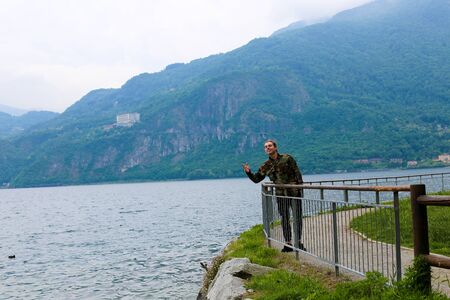 Caucasian tourist wearing camouflage jacket standing near banister,lake Como and mountain in background. Concept of traveling to Italy and tourism.の写真素材