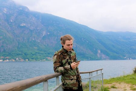 Young male tourist wearing camouflage jacket using smartphone near banister, lake Como and Alps mountain in background. Concept of tourism, modern technology.の写真素材