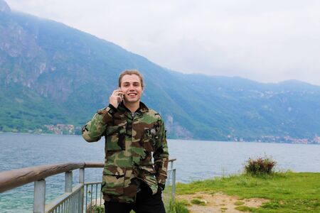 Caucasian male tourist wearing camouflage jacket speaking by smartphone near banister, lake Como and Alps mountain in background. Concept oF tourism, modern technology.の写真素材