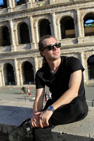 Young man wearng black clothes and sitting near Colosseum background in Rome, Italy. Concept of cheapest tours to Europe and traveling to ancient historic landmarks.の写真素材
