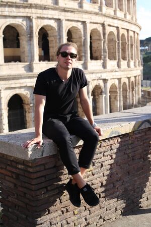 Young caucasian male tourist in glasses sitting on brick wall in Colosseum background in Rome, Italy. Concept of last minute tours to ancient Europe and low cost flights.の写真素材