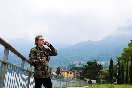 Young european male tourist wearing camouflage jacket speaking by smartphone near banister, lake Como and Alps mountain in background. Concept of Mediterranean sea and tourism, modern technology.の写真素材