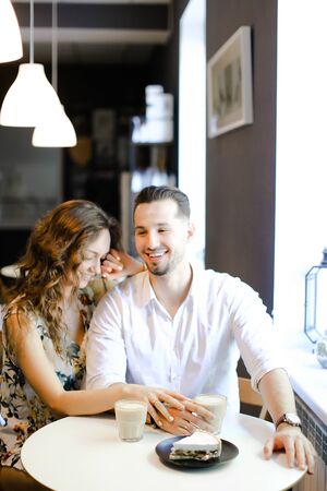 Young smiling boy and girl sitting at cafe with cups of coffe, cake and hugging. Concept of happy couple and taking rest with lunch.の写真素材
