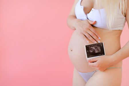 Young happy pregnant woman wearing underwear and holding belly with ultrasound in pink monophonic background. Concept of expectant photo session at studio.の写真素材