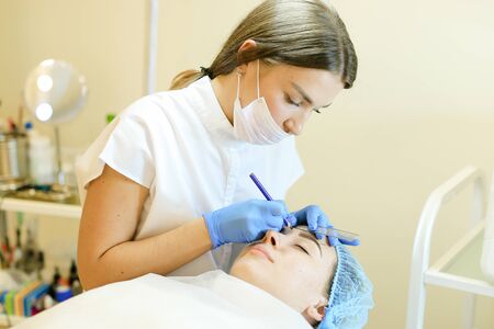 Skilled cosmetologist shaping eyebrows for young caucasian girl before microblading. Concept of permanent makeup artist and beauty salon.の写真素材