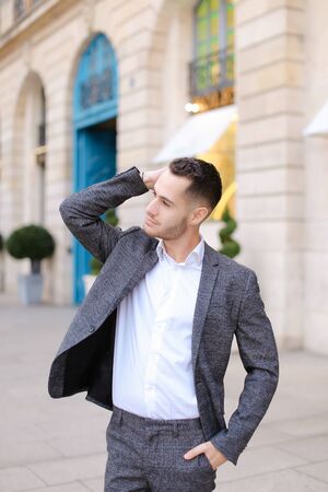 Young cacuasian male person wearing suit standing near building outdoors with rised hand. Concept of fashion and businessman, urban life.の写真素材