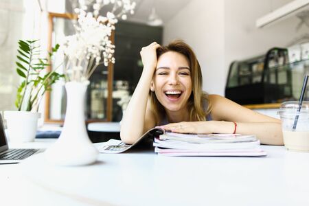 Young smiling woman sittting at cafe with magazines, coffee and laptop. Concept of learning and having break, student life.の写真素材
