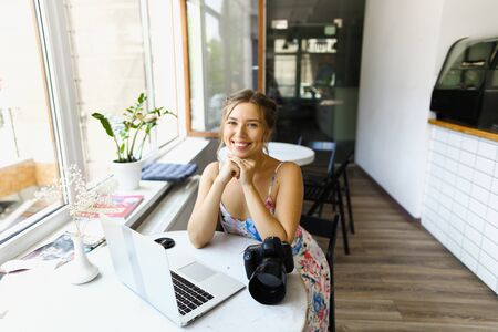 American smiling young female photographer sitting with laptop and camera at cafe. Concept of modern technology and photoartist.の写真素材