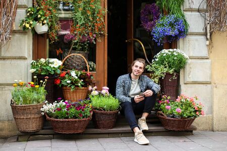 Young man keeping smartphone and sitting at flower shop near bouquets in baskets. Concept of business and modern technology.の写真素材