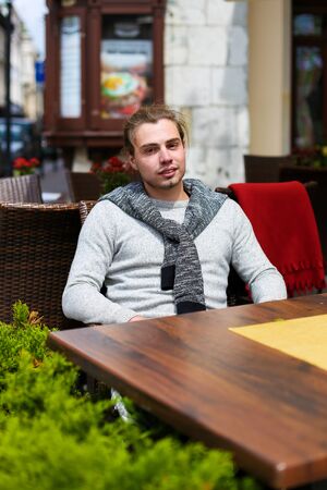 Young man sitting at street cafe near green plants, wearing grey sweater. Concept of resting in open air and waiting lunch.の写真素材