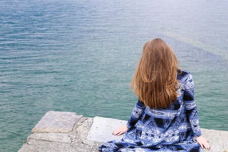Back view of young woman sitting near lake Como, water in background. Concept of summer vacations.の写真素材