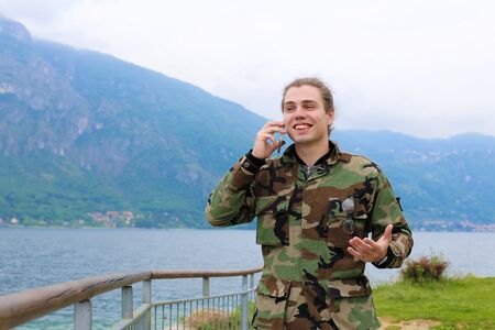 Young happy male tourist wearing camouflage jacket speaking by smartphone near banister, lake Como and Alps mountain in background. Concept of tourism, modern technology.の写真素材