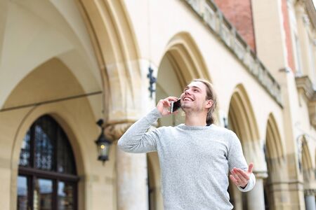 Young caucasian man speaking by smartphone near building with columns in background, wearing grey sweater. Concept of modern technology and visiting Europe.の写真素材