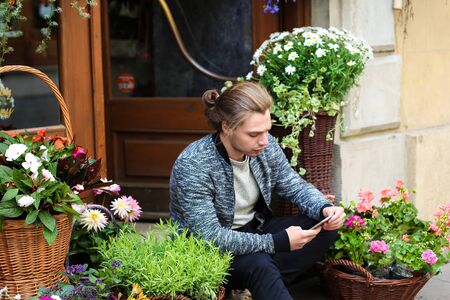 Young boy chatting by smartphone and sitting at flower shop near bouquets in baskets. Concept of business and modern technology.の写真素材