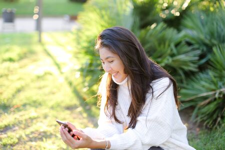 Asian female student sitting in park and typing by smartphone. Concept of modern technology and social networks.の写真素材