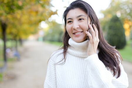 Asian pretty woman wearing white sweater speaking by smartphone walking in autumn park. Concept of chinese girl and modern technology.の写真素材