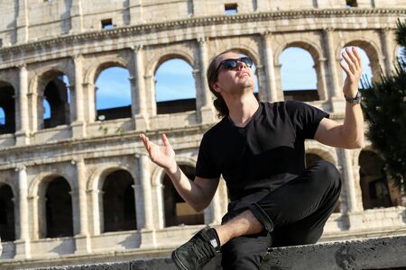 Young male tourist wearng black clothes and sitting near Colosseum background in Rome, Italy. Concept of cheapest tours to Europe and traveling to ancient historic landmarks.の写真素材