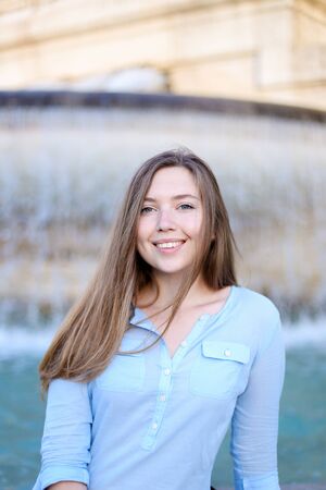 Portrait of young caucasian student sitting near fountain and smiling, wearing blue shirt. Concept of tourism and urban resting.の写真素材