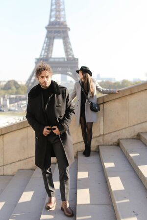 Focus on afro american man in black coat with curly hair on concrete stairs, girl and Eiffel Tower in background, Paris.の写真素材