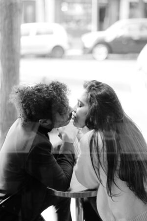 Black and white photo of afro american man kissing caucasian woman and sitting at cafe, reflection in glass. Concept of love and international couple.の写真素材