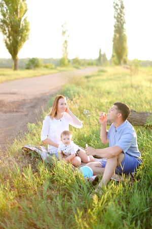 Young father and blonde nice mother sitting on grass with little baby and blowing bubbles. Concept of picnic with child and resting on nature.の写真素材