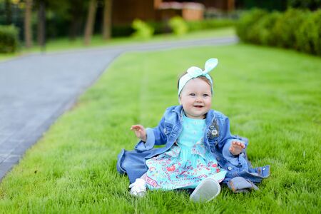 Little cute female baby sitting on green grass and wearing jeans jacket. Concept of childhood and resting in garden, nature background.の写真素材
