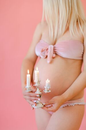 Blonde happy young pregnant woman in underwear holding belly and keeping candles in pink monophonic background. Concept of expactant photo session.の写真素材