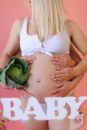 Closeup pregnant woman in underwear keeping inscription baby and cabbage in pink monophonic background. Concept of expactant photo session.の写真素材