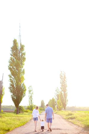 Back view of young mother and father walking on road with little baby, sunshine weather. Concept of parents and children, resting on nature.の写真素材