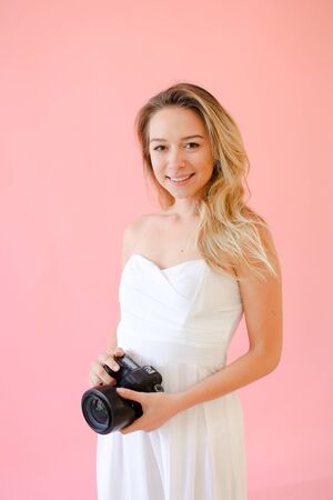 Young european smiling female photographer with camera in pink monophonic background. Concept of modern profession and photo session.の写真素材