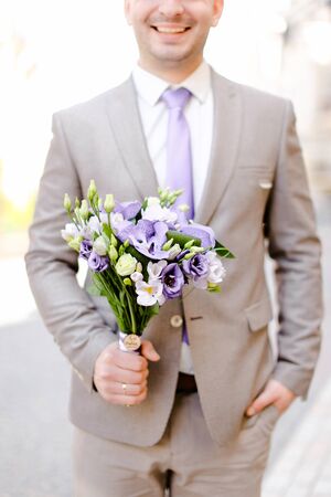 Young caucasian groom keeping bouquet of flowers and wearing grey suit. Concept of wedding photo session and waiting for bride.の写真素材