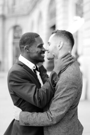 Black and white photo of happy afro american gay hugging caucasian boy outside, wearing suits. Concept of lgbt and same sex couple.の写真素材