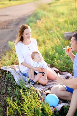 Young mother and father sittling on grass with little baby and blowing bubbles. Concept of picnic and children, parenthood and leisure time.の写真素材