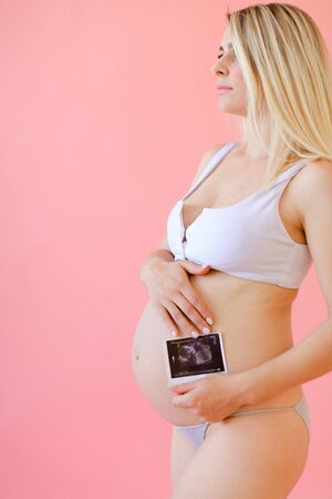 Young happy pregnant woman wearing underwear and holding belly with ultrasound in pink monophonic background. Concept of expectant photo session at studio.の写真素材