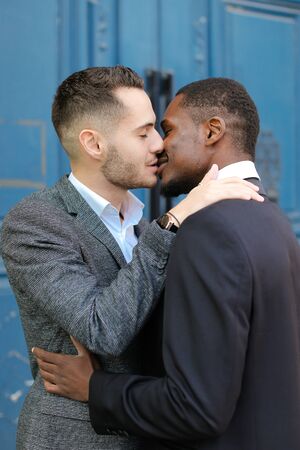Caucasian happy man kissing afro american guy in door background. Concept of lgbt, gays and same sex couple.の写真素材