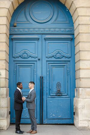Afro american smiling man holding hands of caucasian guy in blue door background, wearing suit. Concept of same sex couple and gays.の写真素材