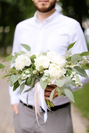 Caucasian groom wearing grey trousers and white shirt waiting for bride with bouquet of flowers. Concept of wedding photo session and floristic art.の写真素材
