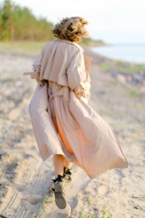 Back view of caucasian young girl walking on shingle beach and wearing summer coat. Concept of seasonal fashion and summer vacations.の写真素材