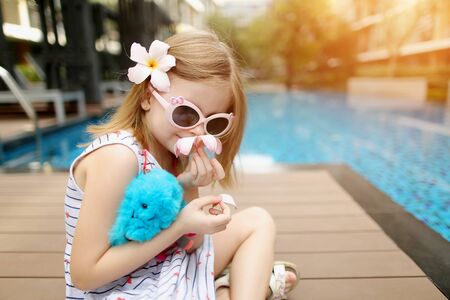 close up portrait of little kid girl sitting close to blue swimming pool wearing sunglasses and hold toy. concept of travel, recreation or holiday family time on sunny dayの写真素材