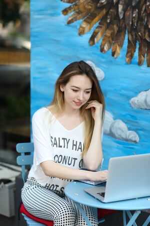 Young female student working with laptop at street cafe near blue painted wall. Concept of modern technology and freelancer worker.の写真素材