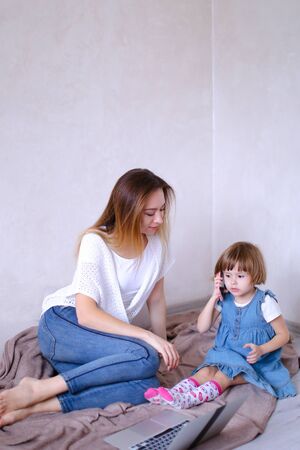 Young mother sitting on bed with little daughter near laptop. Concept of playing with kids, leisure time and modern technology.の写真素材