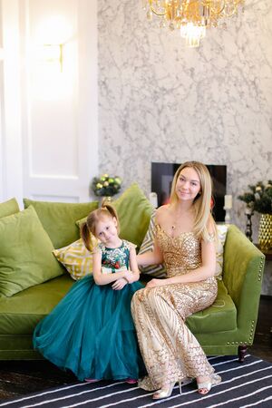 Young mother and little daughter wearing dress sitting on sofa in living room. Concept of celebrating winter holidays and fashionable look.の写真素材