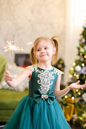 Little beautiful girl wearing dress and standing with bengal lights near Christmas tree. Concept of celebrating winter holidays and childhood.の写真素材