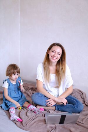 Young smiling mother sitting on bed with little daughter near laptop. Concept of playing with kids, leisure time and modern technology.の写真素材