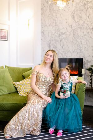 Young caucasian mother and little daughter wearing dress sitting on sofa in living room. Concept of celebrating winter holidays and fashionable look.の写真素材