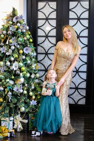 Young caucasian woman and little daughter with gift wearing dresses standing near Christmas tree. Concept of celebrating winter holidays, New Year and fashionable look.の写真素材