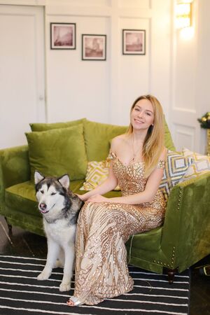 Young woman wearing dress sitting on sofa in living room with husky. Concept of pets, celebrating holidays, fashion and interior.の写真素材