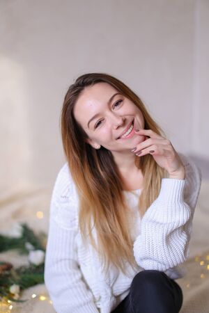 Young european woman sitting on bed near wreath and yellow garlands. Concept of handmade decorations for Christmas and winter holidays.の写真素材