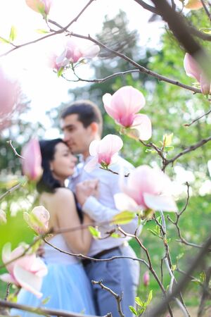 Brunette male person hugging and kissing caucasian girl near magnolia blooming tree. Concept of love and spring season.の写真素材