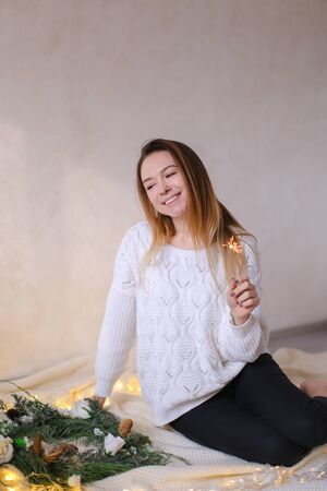 Young cute woman sitting with bengal light near twinkling yellow garlands and green wreath. Concept of handmade decorations for Christmas and New Year holidays.の写真素材